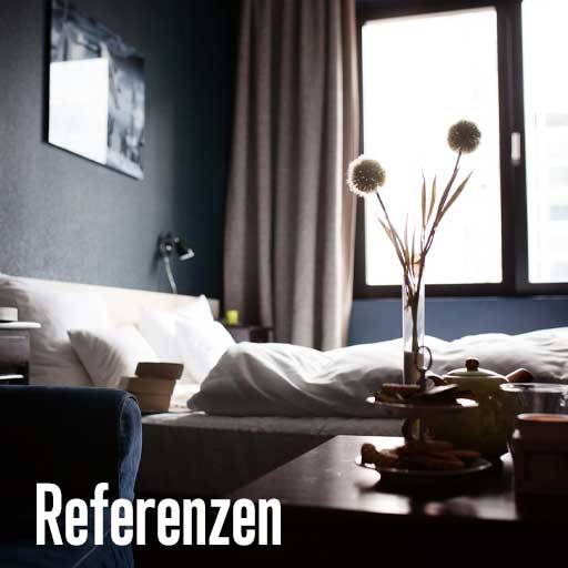 Ein Hotelschlafzimmer, im Fokus ein Bett mit einem Kissen, einer Vase mit Trockenblumen