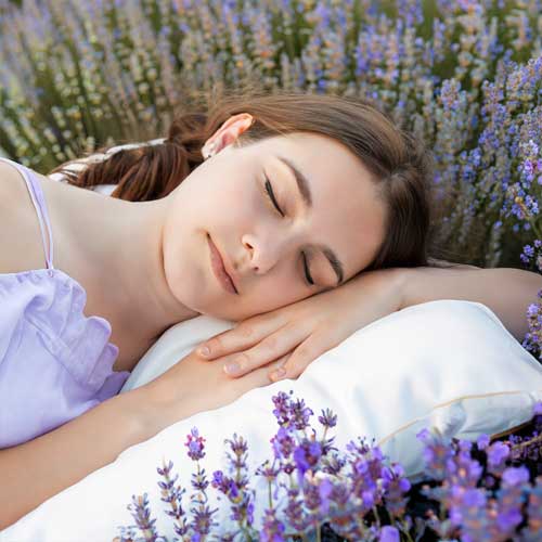 Eine junge Frau schläft entspannt auf einem Lavendel-Kissen, umgeben von Lavendelblüten.
